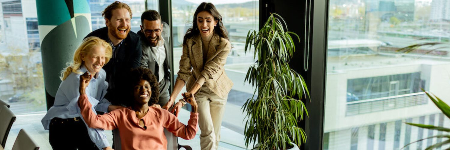 Team of coworkers shares a moment of fun, racing an office chair amidst a well-lit workspace.