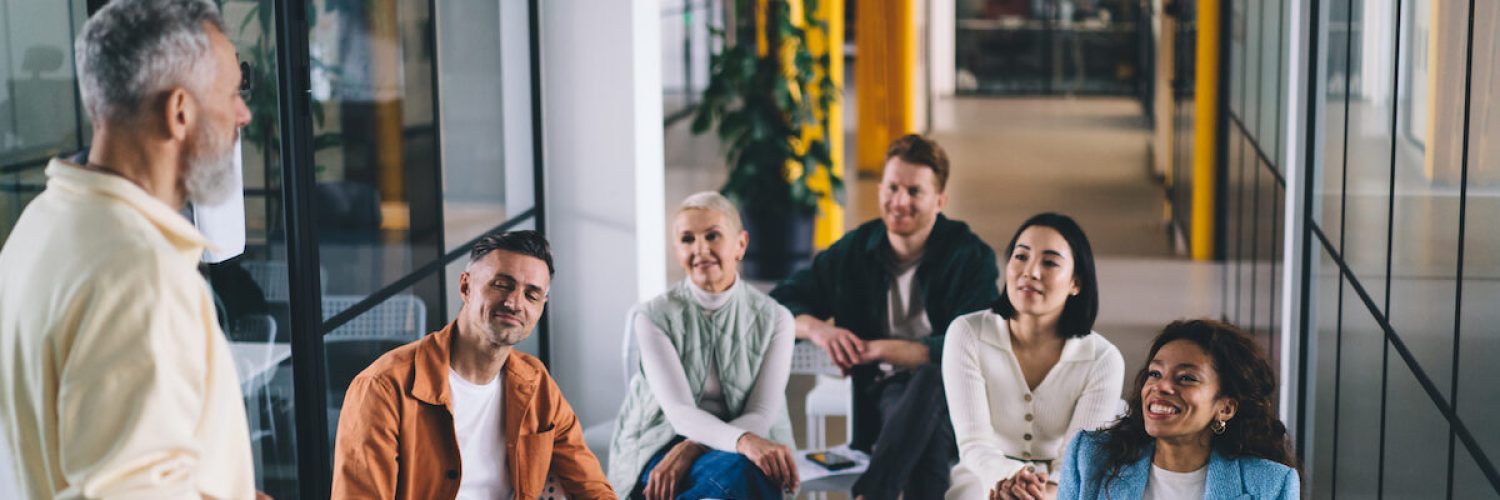 Positive man teaching multiethnic colleagues while standing near glass wall and discussing details of ideas for new startup project in modern workspace at office