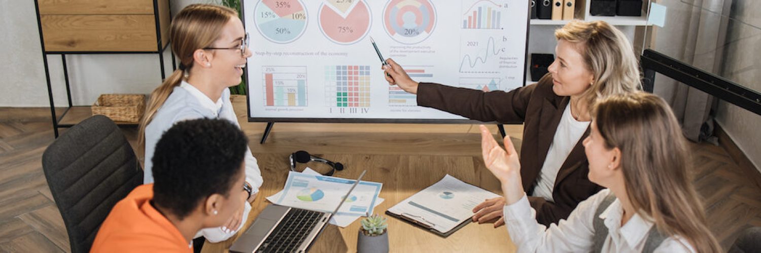 Group of competent female financiers having working meeting at office. Women partners examining report of company. Focus on big computer monitor with statistics graphs.