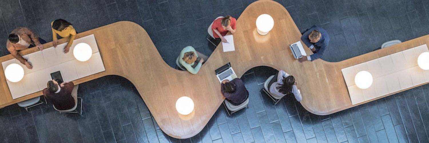 Panoramic overhead view of several business meetings going on in the communal area of a modern office building.